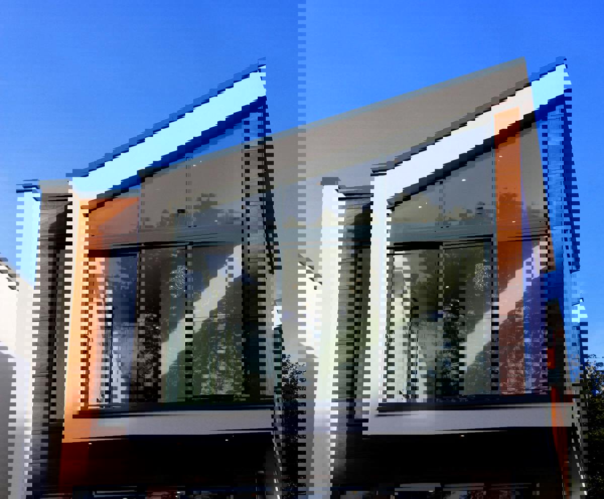 Modern tvåplans villa med stora glaspartier, träpanel och vit putsfasad mot blå himmel.