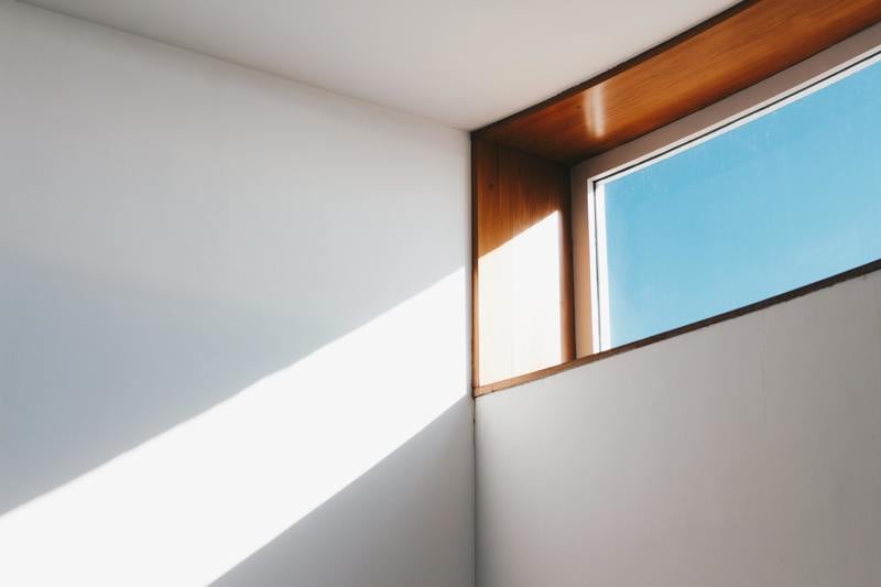 Solljus som lyser in genom ett modernt fönster med träram mot vit vägg och blå himmel utanför.