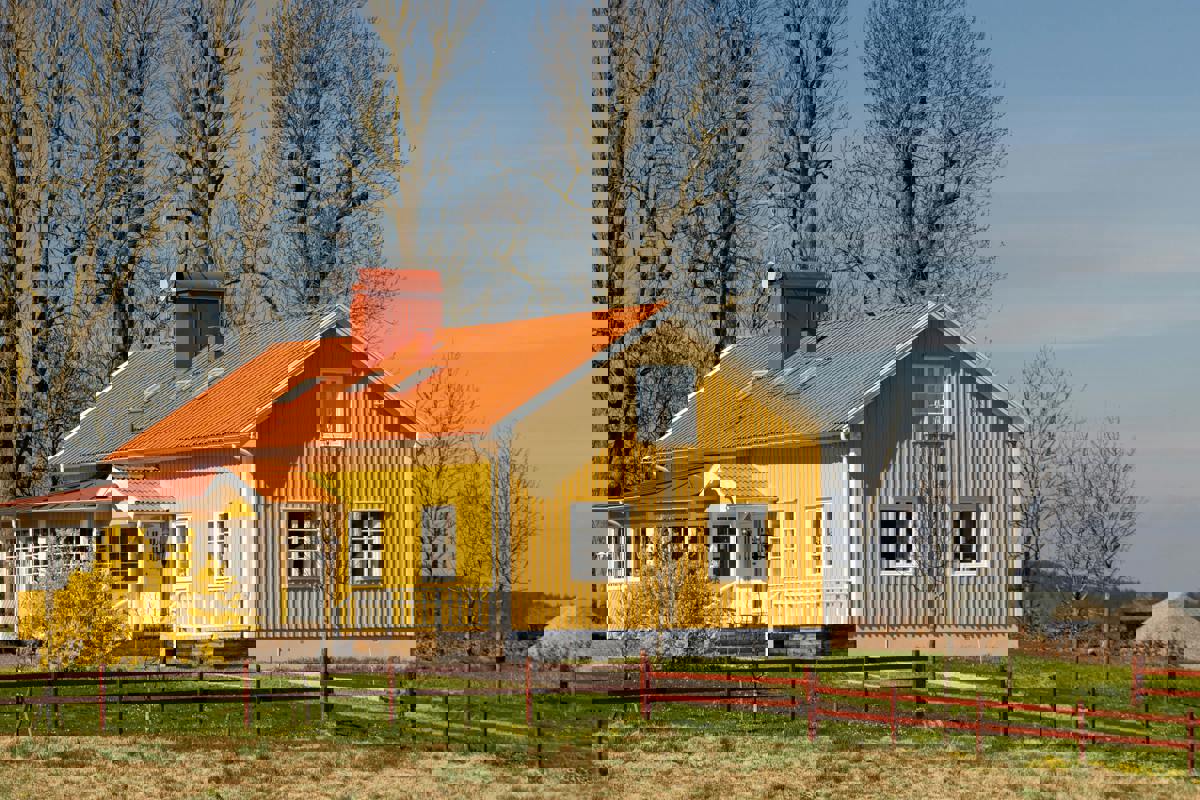 Traditionellt svenskt hus målat i gult med vita knutar och rött tak. Huset står i en lantlig miljö med gräsmatta, träd och blommande buskar i förgrunden, och blå himmel i bakgrunden.