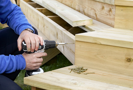 Person skruvar trätrappa med borrmaskin; skruvar ligger bredvid.
