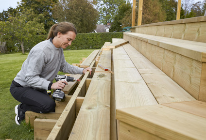 Person skruvar fast träplankor på en trappa i trädgården, med en borrmaskin i handen och skruvar bredvid.