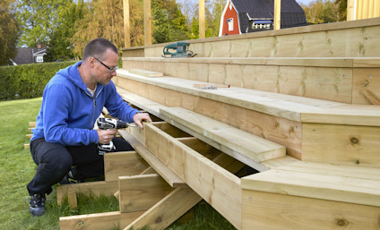 Person som bygger en trappa av trä med hjälp av elverktyg i en trädgårdsmiljö.