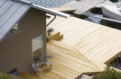 Trädäck med två stolar och bord vid ett hus nära vatten. Brygga och båt synliga i bakgrunden.