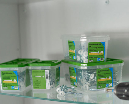 Plastburkar med vita konstruktionsskruvar och gröna etiketter står på en hylla i ett skåp. Övre delen av en svart skruvdragare med en skruv i chucken syns bredvid.