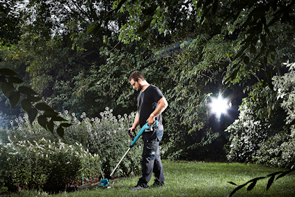 En person trimmar gräs med en batteridriven grästrimmer i en trädgård. Lummiga träd och buskar syns i bakgrunden.