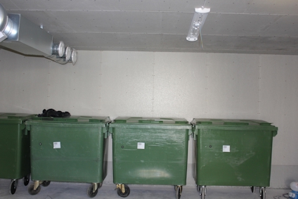 Gröna soptunnor på hjul står uppradade i ett rum med ventilationsrör i taket.