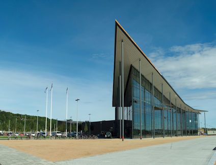 En stor modern byggnad med glasfasad och lutande yttertak, flaggstänger på sidan, klarblå himmel i bakgrunden.