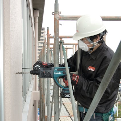 En person i skyddshjälm och jacka använder en Makita-borrmaskin vid ett fönster, omgiven av byggnadsställningar.