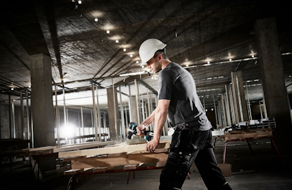 Person med skyddshjälm borrar i trä på en byggarbetsplats med betongtak och pelare.