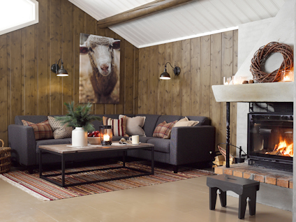 Vardagsrum med braskamin, mörkgrå soffa, bord, fåtölj, vägg med fårbild och vägglampor. Golvmatta i rött mönster.