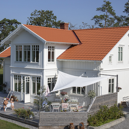 Vit tvåvåningsvilla med rött tegeltak, stor uteplats med solskydd och omgiven av grönska och träd.