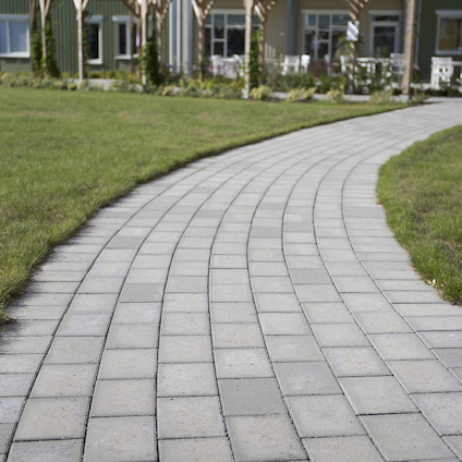 En stenlagd gångväg med grå rektangulära plattor leder mot en byggnad i bakgrunden. Gräs på båda sidor.