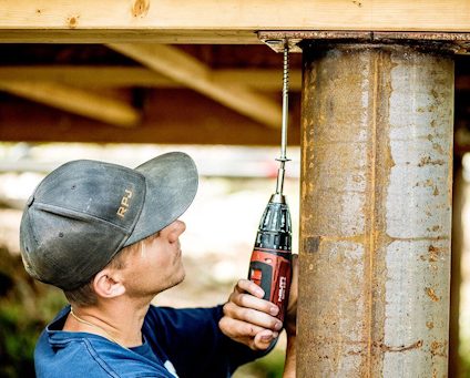 En person monterar en träbjälke mot en stålpelare med en skruvdragare.