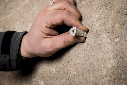 En hand i svart jacka håller en vit lättbetongplugg mot en betongvägg.