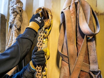 Hand med handske greppar rostig kedja bredvid upphängda bälten och rep mot beige vägg.