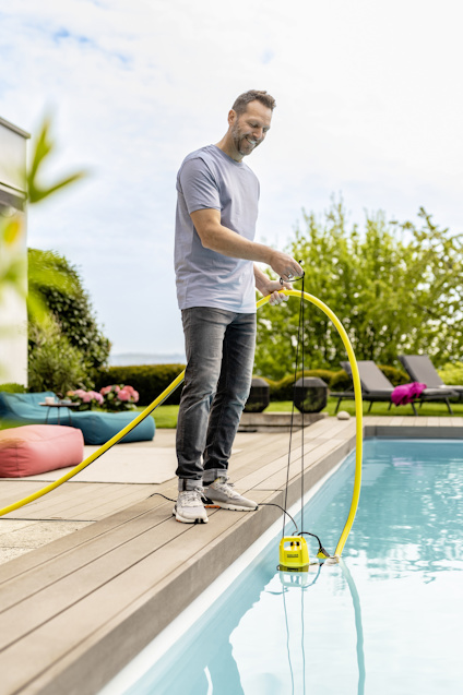 Man kontrollerar en vattenpump med en gul slang vid en pool. Solstolar och gröna träd i bakgrunden.