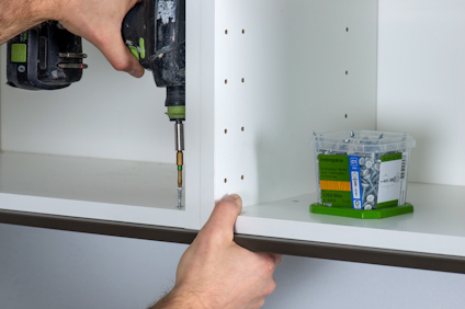 En person skruvar fast en vit hylla med en skruvdragare. Bredvid står en plastlåda med skruvar.