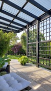 Pergola med träribbor och tak av glas, omgiven av grönska och blommor i trädgårdsmiljö.