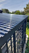 Svart pergola med genomskinligt tak av korrugerad plast, omgiven av grönska och blå himmel.