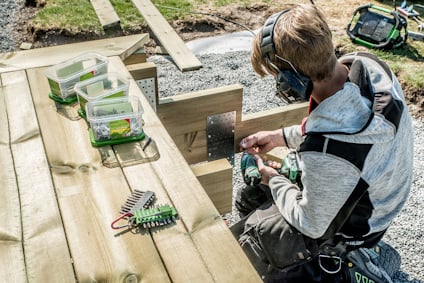 Person bygger med träplankor utomhus, med skruv och borrmaskin. lövträ produkter och skyddsutrustning syns.