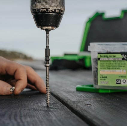 Skruvdragare med bit och skruv, hand i bakgrunden, låda med däckskruvar på ett träbord utomhus.