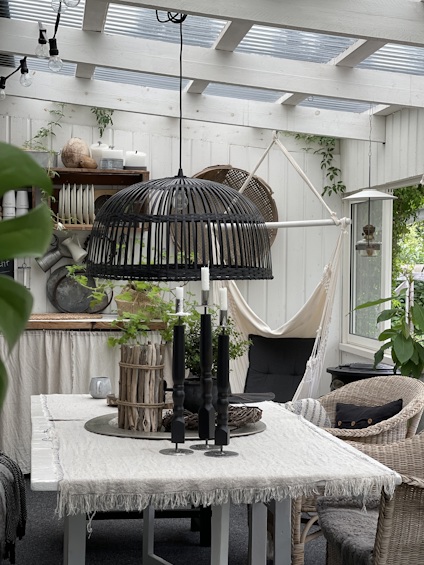 Inomhus veranda med korgmöbler, svart korglampa, växter och bord med duk och ljusstakar.