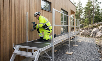 En person i varselkläder arbetar på en byggnadsställning intill en trävägg.